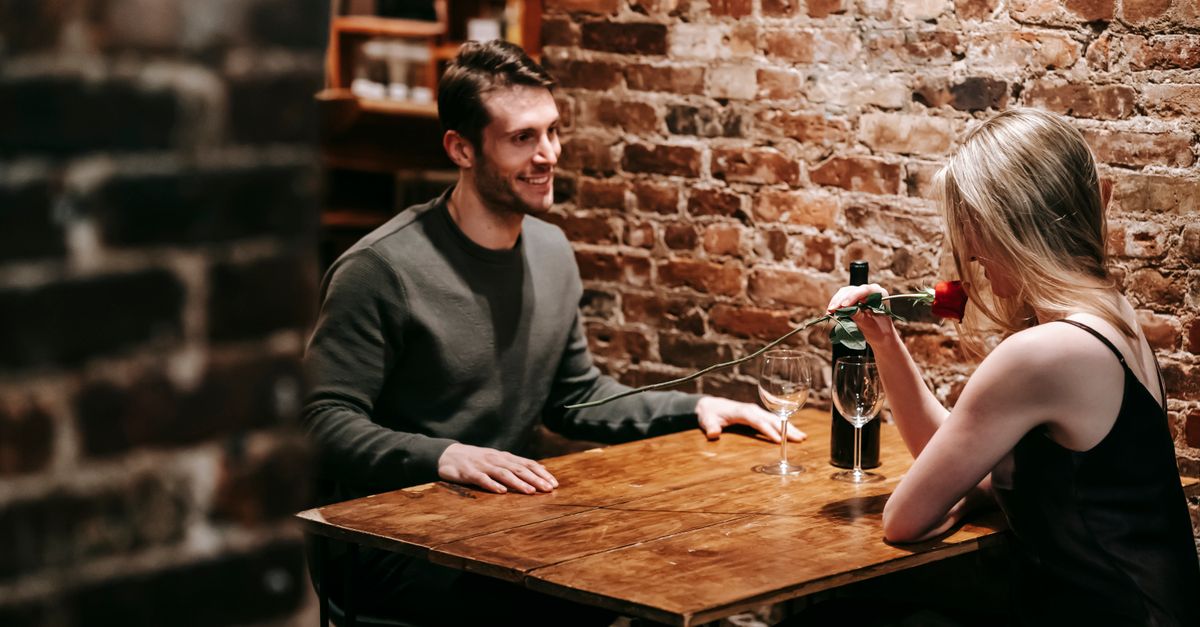 Young couple in elegant clothes sitting in light cafeteria at wooden table near brick wall and having romantic dinner with red rose and wine near glasses
